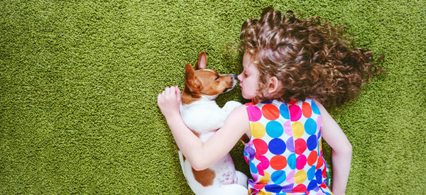 little girl in spotty coloured dress laying on green carpet with small dog in arm